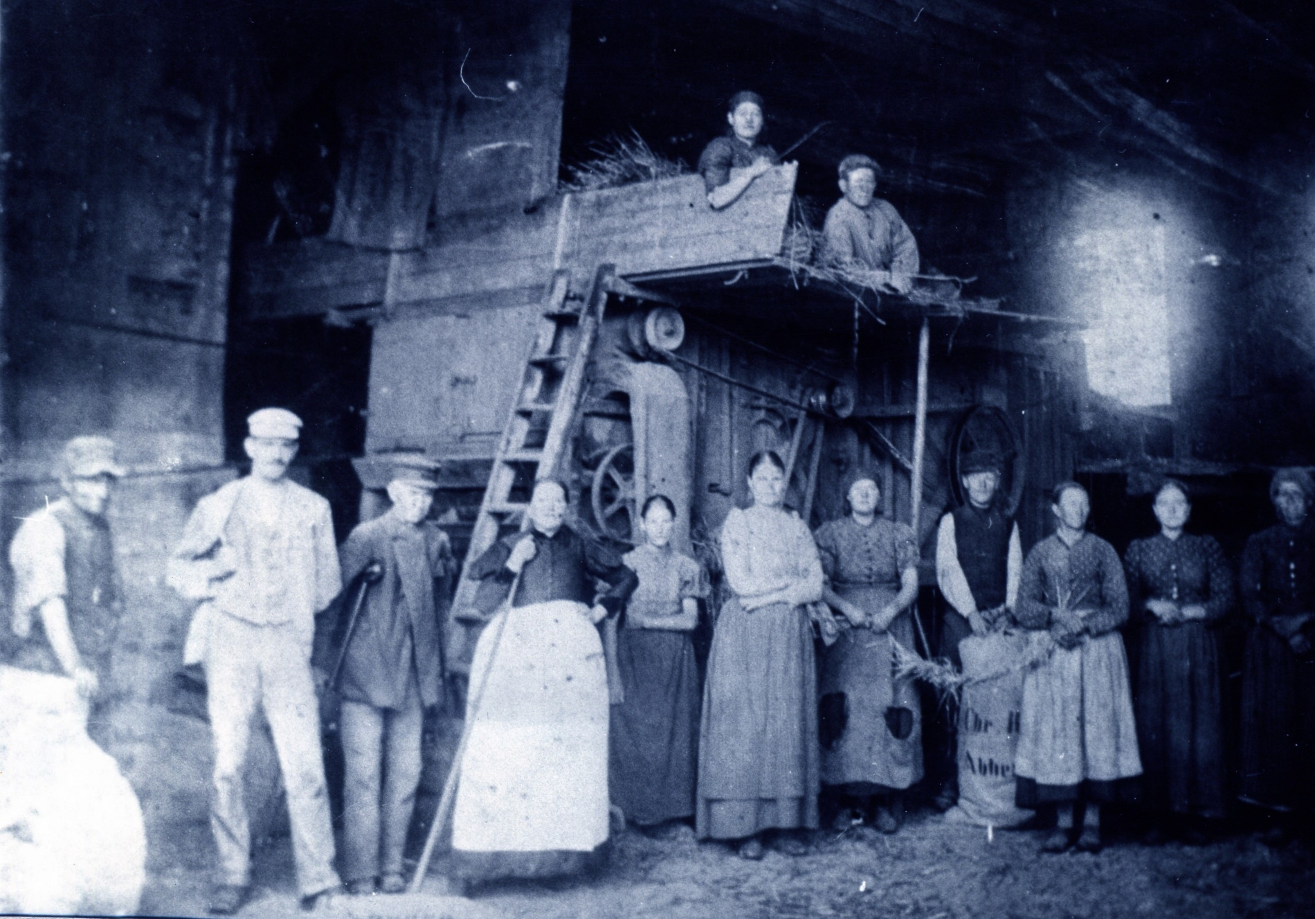 Historische Aufnahme in schwarz-weiß. 11 Männer und Frauen stehen in einer Reihe in einer Art Scheune und blicken zur Kamera. Zwei weitere Personen liegen darüber auf einer Art Heuboden. Es sind historische Gerätschaften für den Mühlenbetrieb zu erahnen.