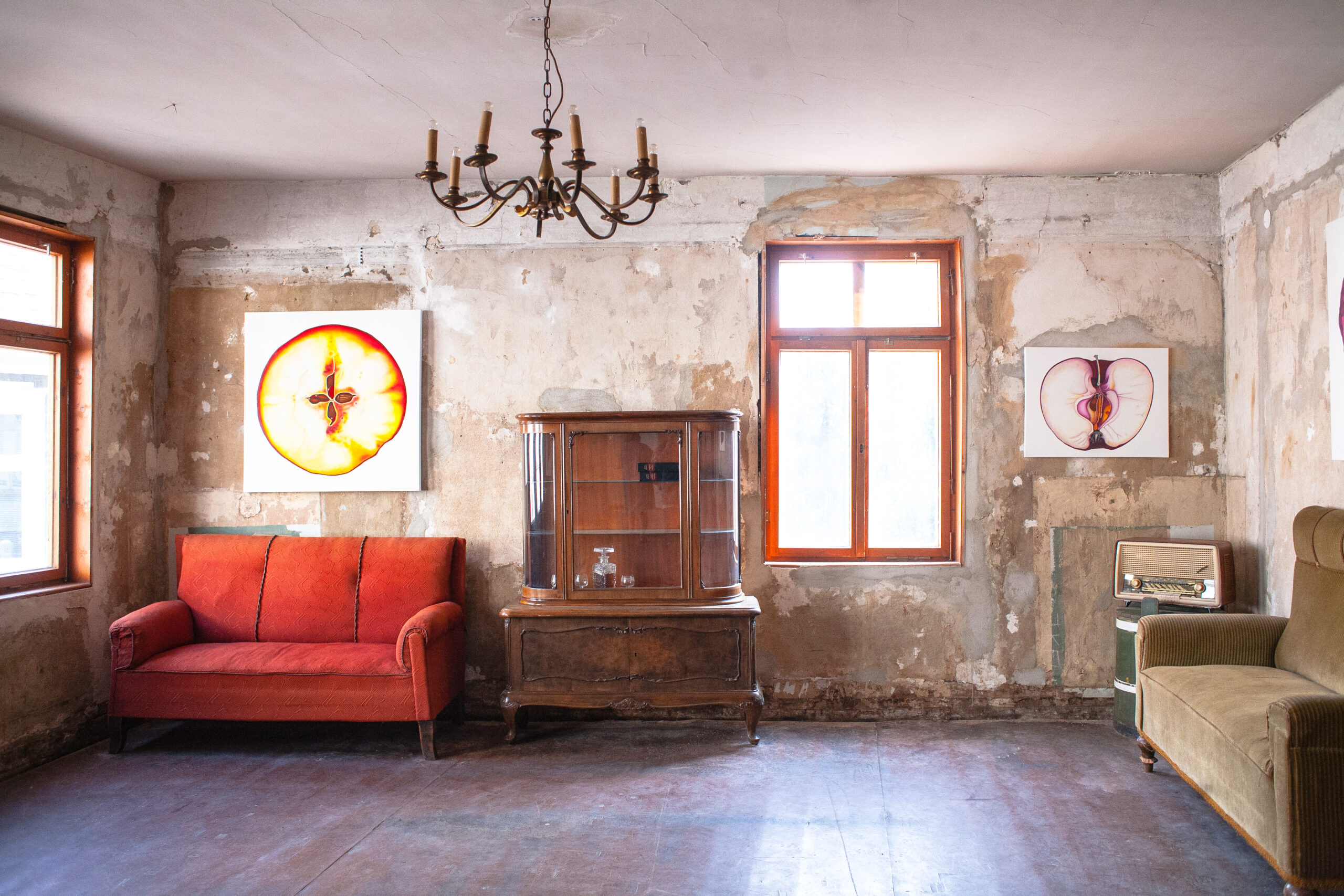 Blick in einen Raum mit einer unverputzten Wand und einem dunklen Holzboden. Links hinten steht ein rotes Sofa, daneben eine alte Holzvitrine. An der Decke hängt ein Kronleuchter. Es hängen zwei künstlerische Darstellungen eines Apfels an der Wand.