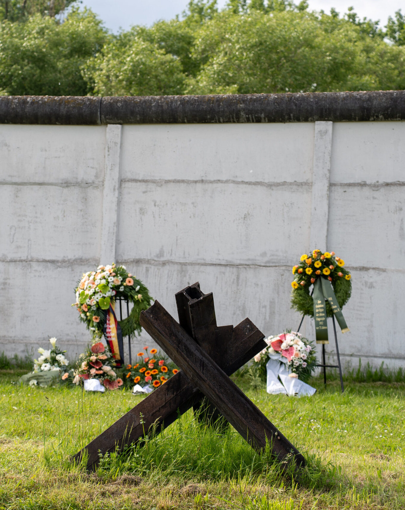 Vor der Grenzmauer liegen mehrere Blumenkränze mit Schleifen. Im Vordergrund eine Panzersperre.