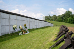 Blick entlang der Grenzmauer, vor der einige Blumenkränze liegen. Rechts verläuft eine Reihe von Panzersperren, im Hintergrund ein Grenzturm.