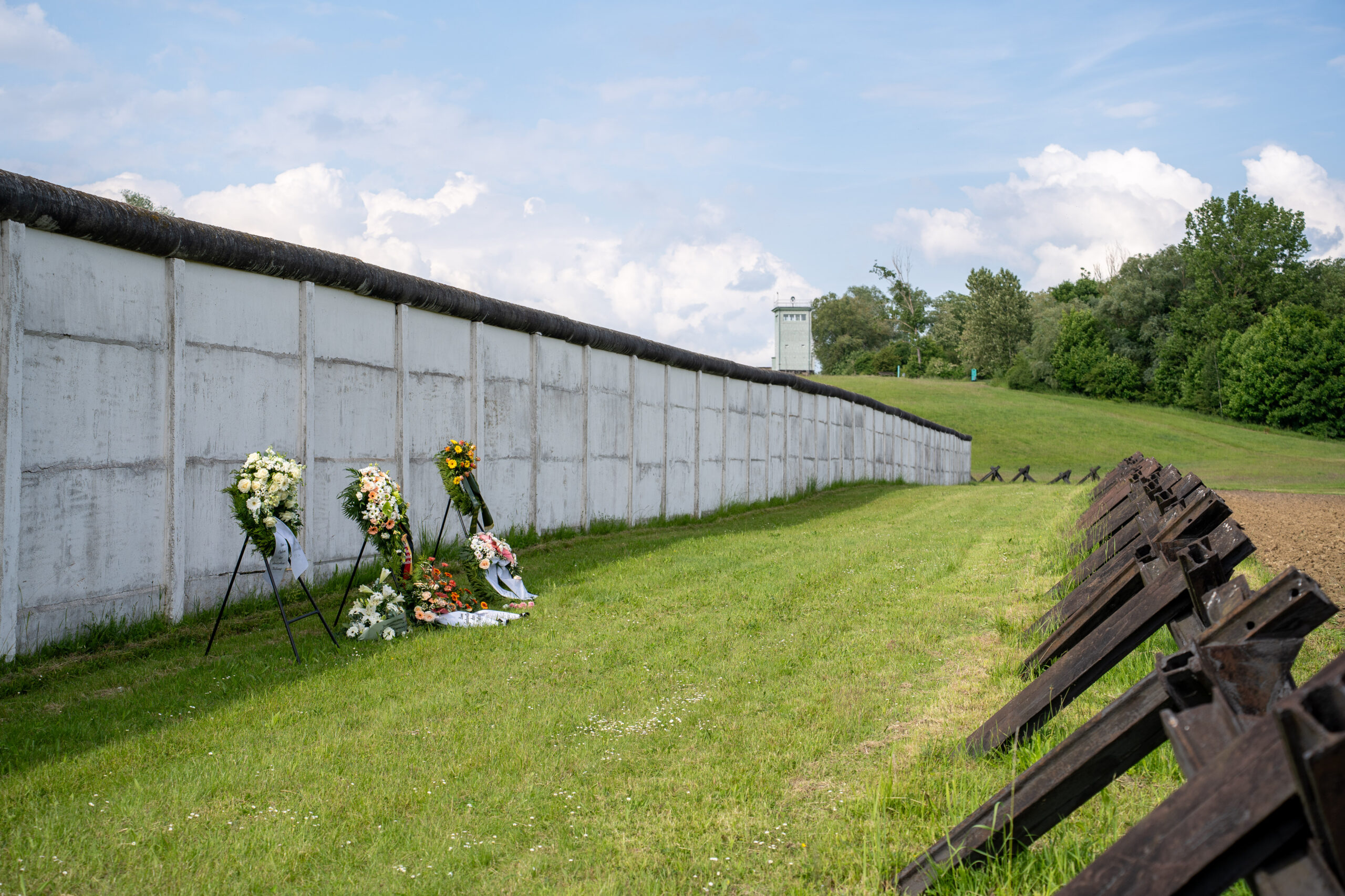 Blick entlang der Grenzmauer, vor der einige Blumenkränze liegen. Rechts verläuft eine Reihe von Panzersperren, im Hintergrund ein Grenzturm.