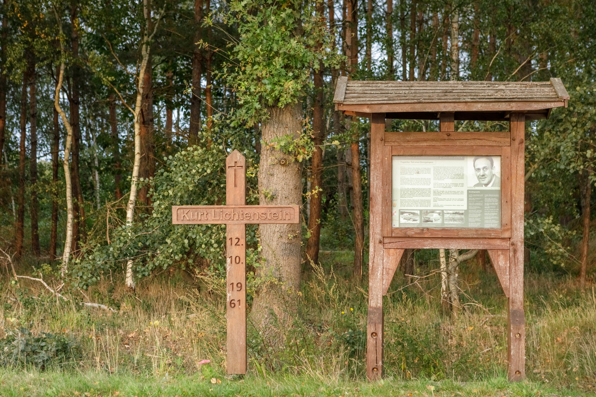 Holzkreuz mit der Aufschrift "Kurt Lichtenstein. 12.10.1961" und daneben eine Gedenktafel mit Text und einem Foto. Im Hintergrund Wald.